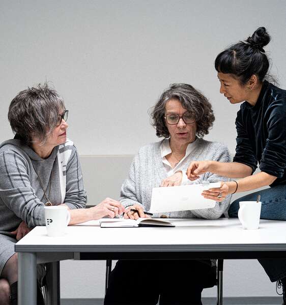 Drei Frauen sitzen an einem Tisch mit einem Notizbuch und einem Tablet. Eine Frau zeigt auf das Tablet, während die anderen beiden aufmerksam schauen. Auf dem Tisch stehen zwei Tassen.