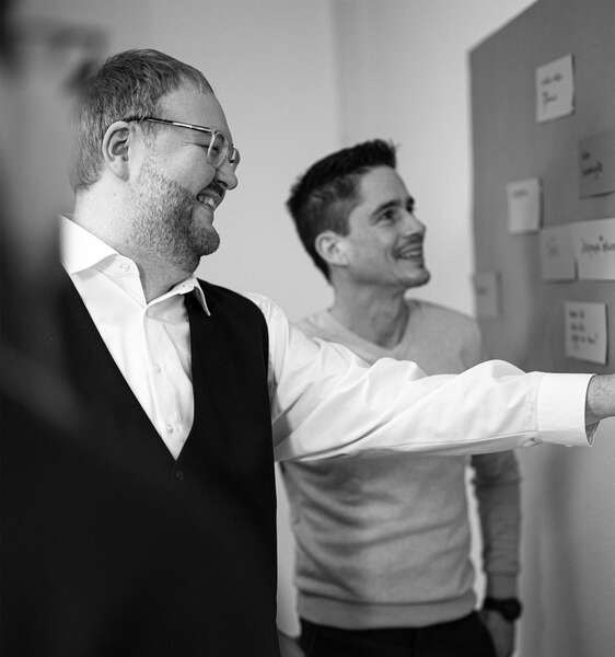 Schwarz-Weiß-Foto: Ein Mann in einem weißen Hemd mit einer schwarzen Weste zeigt auf eine Wand mit Notizen. Ein weiterer Mann in einem grauen Pullover steht daneben.