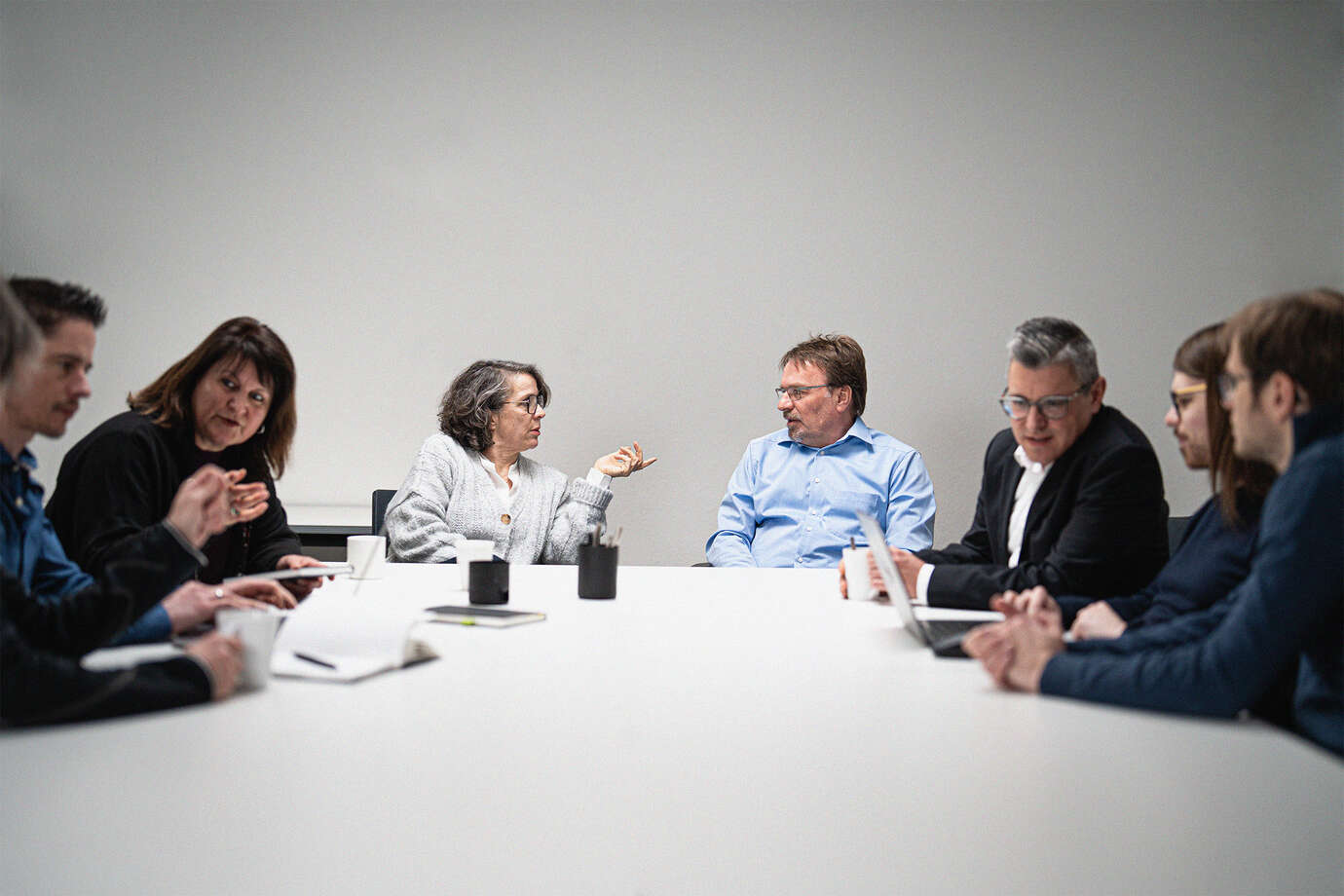 Eine Gruppe von sieben Personen sitzt an einem Tisch in einem Besprechungsraum. Eine Person in der Mitte gestikuliert, während andere zuhören. Auf dem Tisch liegen Notizblöcke und Stifte.