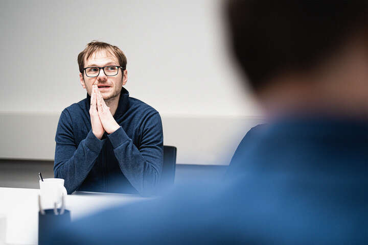 Ein Mann sitzt an einem Tisch mit gefalteten Händen. Er trägt einen blauen Pullover und blickt nachdenklich. Im Vordergrund ist eine weitere Person unscharf zu sehen.