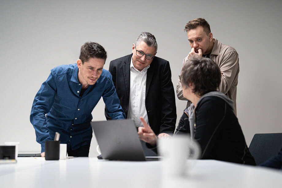 Vier Personen stehen um einen Tisch und betrachten einen Laptop. Zwei Personen tragen Hemden, eine Person einen Anzug und eine Person einen Pullover.
