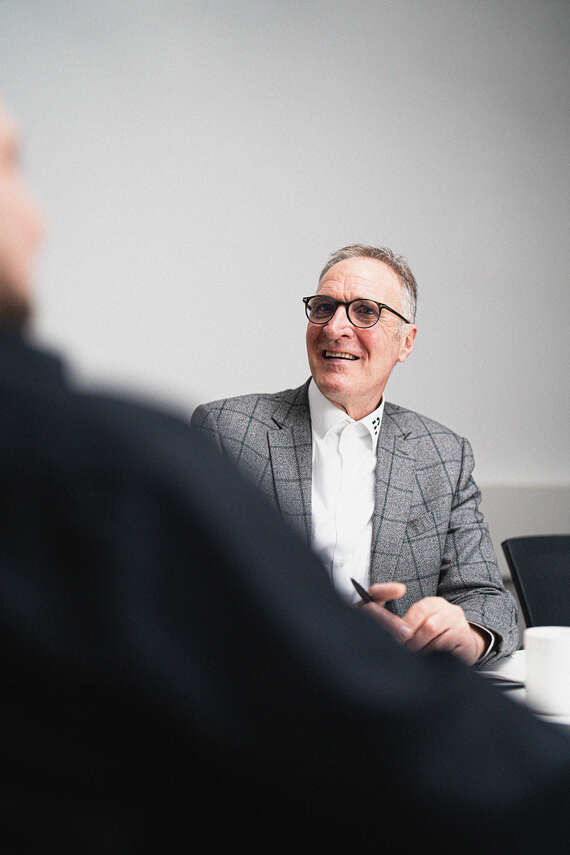 Ein Mann in einem karierten Anzug und Brille sitzt an einem Tisch und hält einen Stift in der Hand. Eine Tasse steht auf dem Tisch.