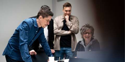 Drei Personen in einem Raum, eine lehnt sich nach vorne auf einen Tisch, eine steht mit Hand am Kinn, eine sitzt vor einem Laptop.