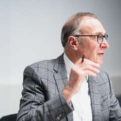 Älterer Mann in grauem Karo-Sakko und weißem Hemd hebt die rechte Hand, Blick nach rechts gewandt.