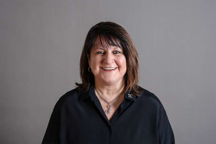 Frau mit braunen, schulterlangen Haaren trägt schwarze Strickjacke und lila Oberteil, sitzt an einem Tisch.