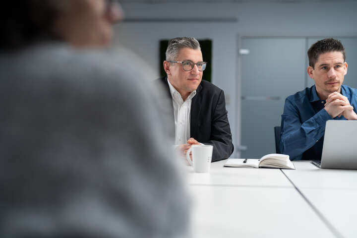 Zwei Männer sitzen an einem weißen Tisch mit Notizbuch und Kaffeetasse, im Vordergrund eine unscharfe Person.