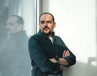 Teammitglied mit kurzen Haaren und Bart in schwarzem Oberteil mit verschränkten Armen, selbstbewusste Pose vor hellem Hintergrund im Portrait.