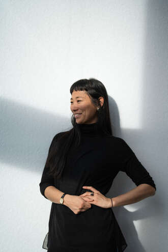 Teammitglied mit schulterlangen Haaren in schwarzem Rollkragenpullover, nachdenkliche Pose mit verschränkten Händen vor hellem Hintergrund im Portrait.