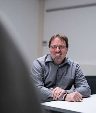 Teammitglied mit Brille und Bart in grauem Pullover, freundlich lächelnd an einem Bürotisch sitzend in heller Büroumgebung im Portrait.