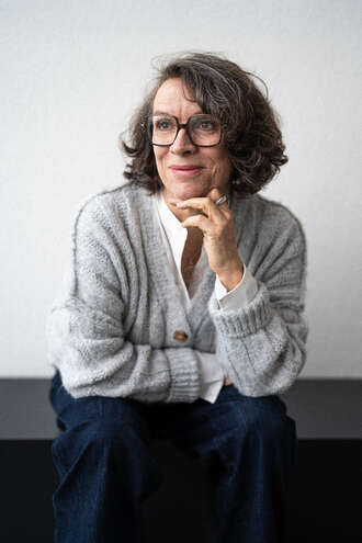 Teammitglied mit Brille und schulterlangen Haaren in grauer Strickjacke und dunklem Rock, nachdenkliche Pose mit Hand am Kinn im Schwarz-Weiß-Portrait.