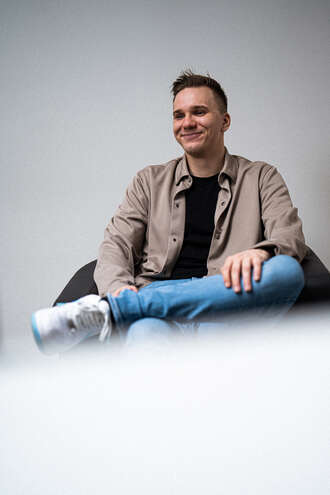 Teammitglied mit kurzen Haaren in beiger Jacke und Jeans, lächelnd in entspannter Sitzposition mit Dokumentenrolle auf weißem Hintergrund.