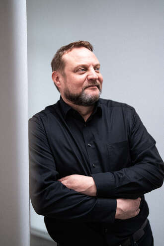 Teammitglied mit Vollbart in schwarzem Hemd mit verschränkten Armen, selbstbewusste Pose mit Blick zur Seite vor grauem Hintergrund im Business-Portrait.