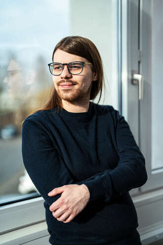 Teammitglied mit Brille und schulterlangen Haaren in schwarzem Pullover, lächelnd mit verschränkten Händen vor hellem Fensterbereich im Business-Portrait.