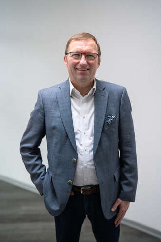 Teammitglied mit Brille in grauem Sakko und weißem Hemd, selbstbewusste Haltung mit Hand in der Hosentasche vor neutralem Hintergrund im Business-Portrait.
