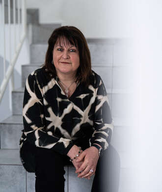 Teammitglied mit schulterlangen dunklen Haaren in schwarz-weißem Muster-Pullover und schwarzer Hose, freundlich lächelnd auf einer weißen Treppe sitzend.