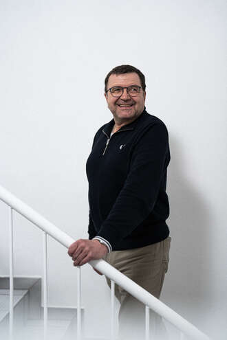 Teammitglied mit Brille in schwarzem Pullover und beiger Hose, lächelnd und mit der Hand am Treppengeländer vor weißer Wand stehend.