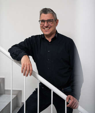 Teammitglied mit Brille und grauem Haar in schwarzem Hemd, entspannt mit einer Hand am weißen Treppengeländer lehnend vor hellem Hintergrund.