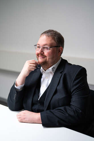Teammitglied mit Brille in dunklem Sakko und weißem Hemd, nachdenklich mit Hand am Kinn an einem Bürotisch sitzend im Business-Portrait.