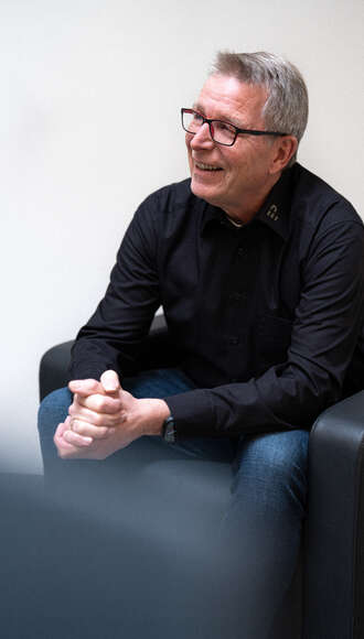 Teammitglied mit Brille und grauen Haaren in schwarzem Hemd, entspannt sitzend mit gefalteten Händen vor weißem Hintergrund im Business-Portrait.