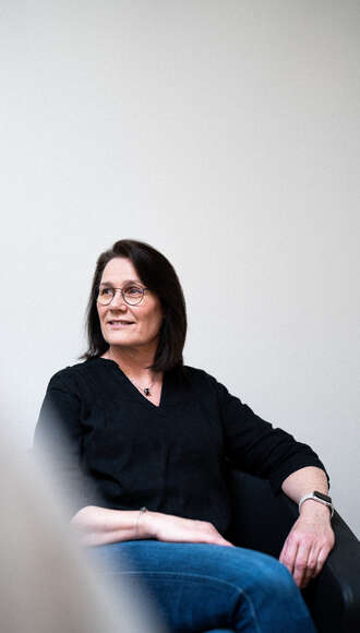 Teammitglied mit Brille und dunklen Haaren in schwarzem Oberteil, seitlich auf weißem Stuhl sitzend mit entspannter Körperhaltung vor hellem Hintergrund.