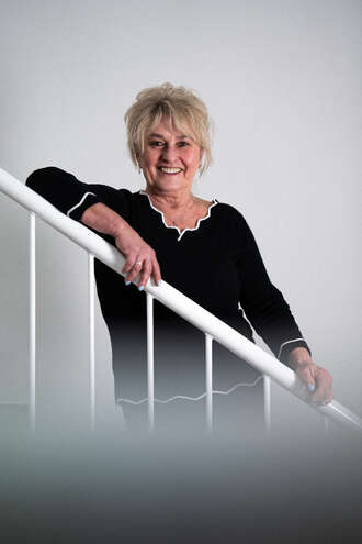 Teammitglied mit Brille und blonden kurzen Haaren in schwarzem Oberteil, freundlich lächelnd an einem weißen Treppengeländer stehend im Business-Portrait.