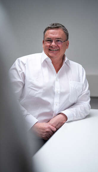 Teammitglied mit grauen Haaren in weißem Hemd, freundlich lächelnd mit verschränkten Armen vor hellem Hintergrund im professionellen Portrait.