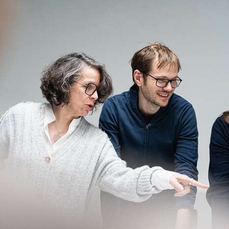 Eine ältere Frau mit grauen, lockigen Haaren trägt einen hellen Pullover und zeigt mit dem Finger auf etwas. Neben ihr steht ein jüngerer Mann mit bräunlichen Haaren in einem blauen Pullover.